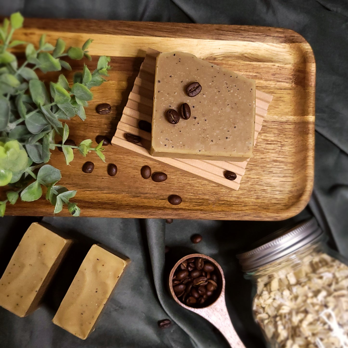 Wooden tray with coffee-scented soap bars and coffee beans, surrounded by a plant and jar of oats.