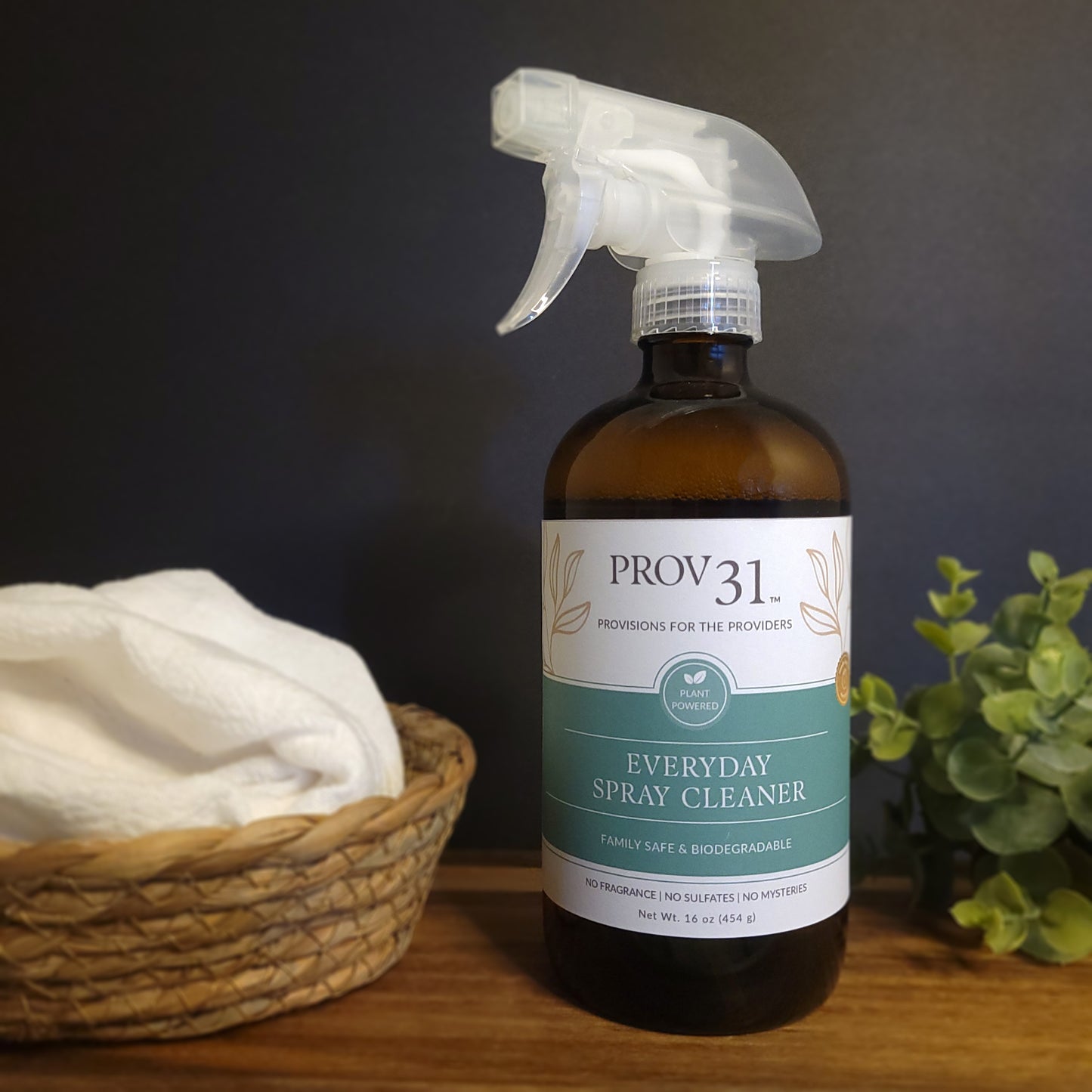Bottle of Prov31 Everyday Spray Cleaner on a wooden surface with a basket and plant in the background.