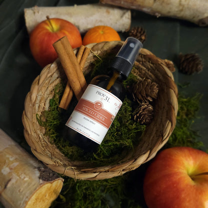 Bottle of Prov31 Spiced Cider Room Spray with cinnamon sticks and apples in a basket.