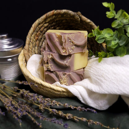 Three bars of purple soap with loofah shavings in a woven basket with lavender and a white cloth.