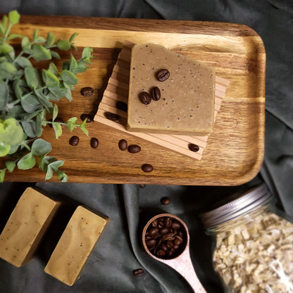 Wooden tray with coffee-scented soap bars and coffee beans, surrounded by a plant and jar of oats.